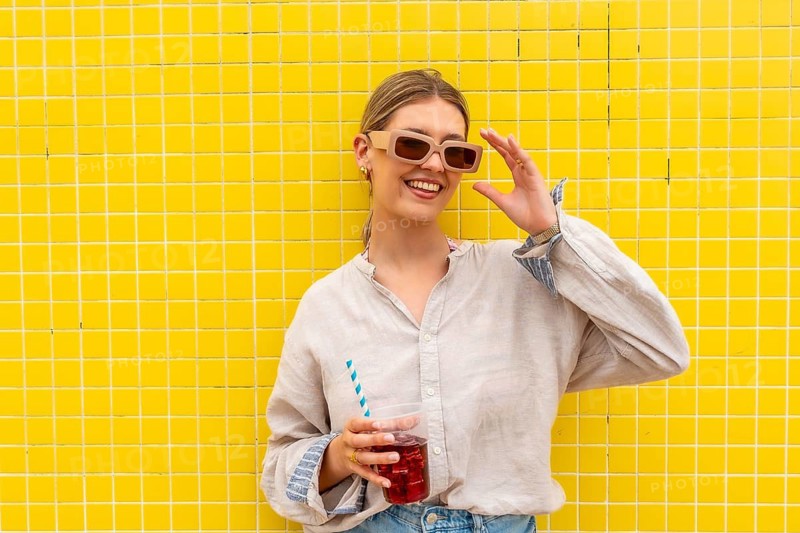 Young woman leaning on a vibrant yellow tiled wall, adjusting her sunglasses with one hand while holding a refreshing drink with a striped straw, smiling and enjoying summer