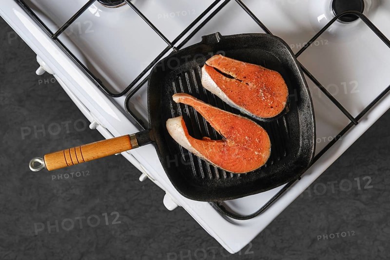 Salmon fillets are placed on cast iron grill pan over gas stove. Fish is seasoned and being cooked indoors. The kitchen has simple design with focus on cooking process