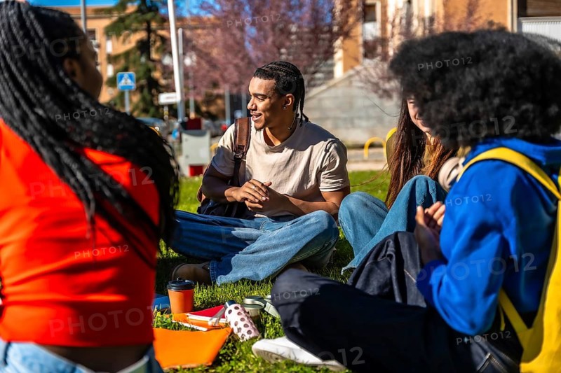 Diverse group of college students sitting on grass at a university campus, chatting, studying and enjoying a sunny outdoor break together with backpacks, books and drinks