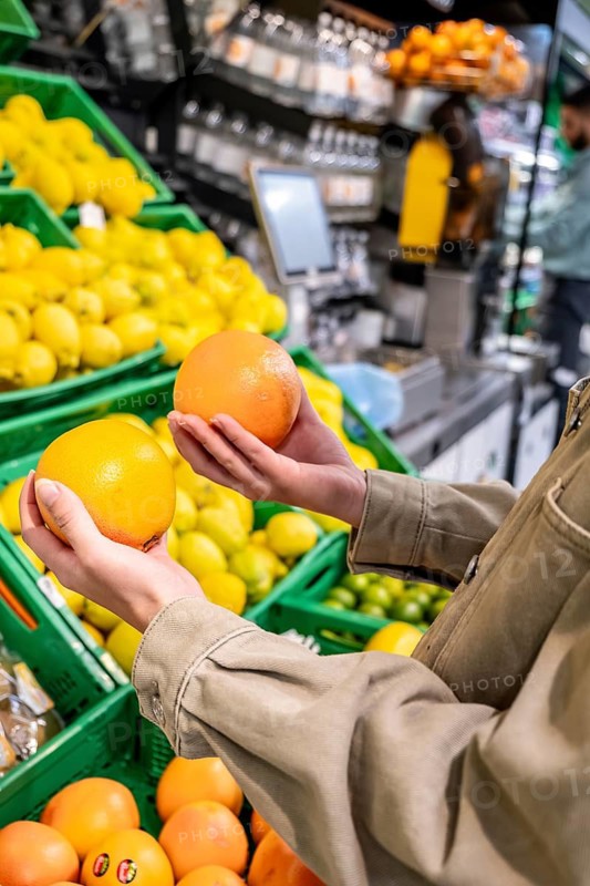 Person's hands holding and comparing two grapefruits in a supermarket produce aisle filled with fresh citrus fruits like lemons and limes, emphasizing healthy food choices
