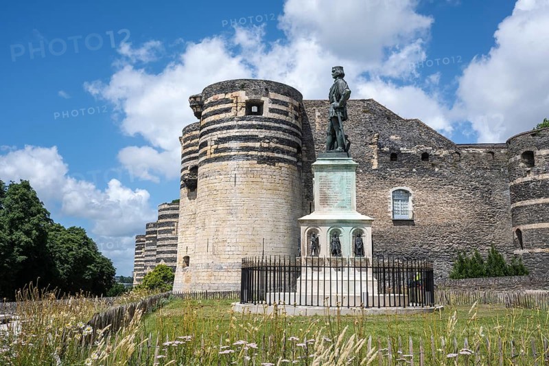 Bronze statue of King René on a decorated stone base in the foreground in front of one of the mighty, striped defensive towers of the historic Château d'Angers castle complex made of slate and limestone under a blue summer sky with loose clouds, Angers, Maine-et-Loire, Pays de la Loire, France