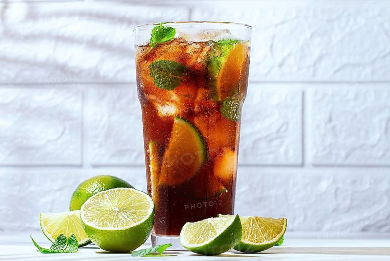 A glass filled with a cola beverage is surrounded by lime slices and fresh mint leaves. Whole limes sit nearby on a marble surface. Sunlight casts shadows