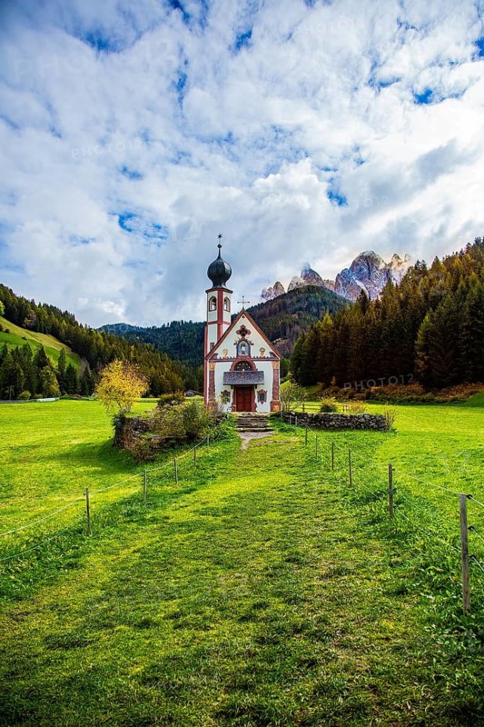 Church in a vast green valley surrounded by mountains and forests with blue skies and clouds, Church of St. Johann in Ranui, Villnöss Valley, South Tyrol, Italy, A narrow path leads to a church against a background of Alps and cloudy sky, St. Johann in Ranui Church with Geisler Group, Villnöss Valley, South Tyrol, Italy