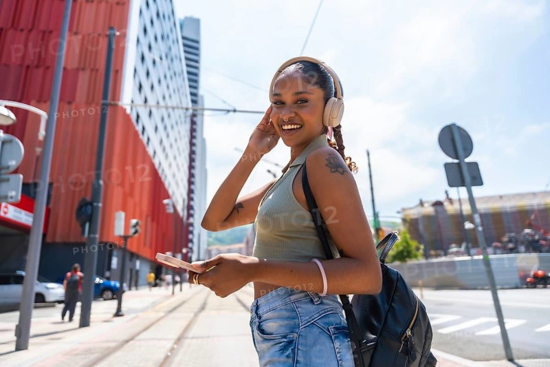 Young woman smiling and wearing headphones, standing on an urban street with a backpack and holding a smartphone, enjoying music in a city environment