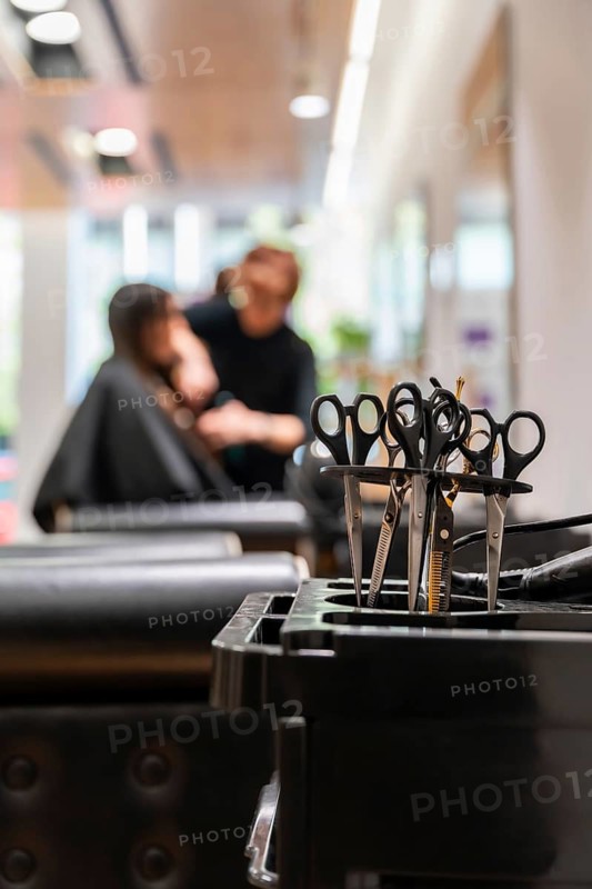 Professional hair cutting scissors standing in a black barber cart as a blurred hairdresser styles a client's hair in the background, showcasing beauty salon work