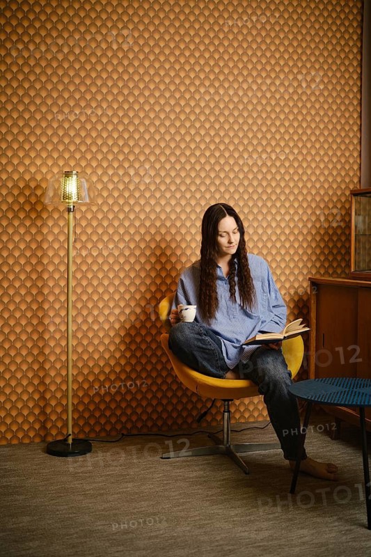 Woman with long hair sits in yellow chair. She holds book in one hand and cup in other. Room has brown patterned wall. Floor lamp and wooden furniture are present in background