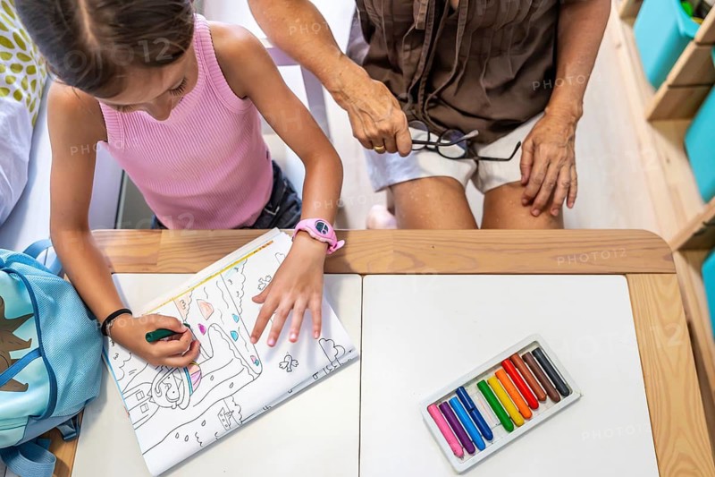 Grandmother and granddaughter bonding, coloring a book at a desk with crayons, enjoying creative time and childhood development activities