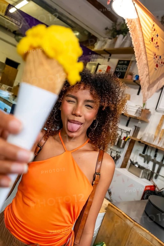 Young woman with curly hair showing a playful expression and sticking out her tongue while holding a mango ice cream cone, enjoying a refreshing treat during summer