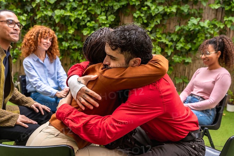 Two diverse individuals embracing during a support group, showing compassion and emotional connection while other multiethnic participants listen in a mental health and recovery setting