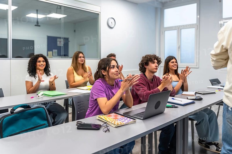 Happy diverse students are sitting at desks, clapping their hands in a modern classroom or university lecture hall, showing appreciation and feeling success