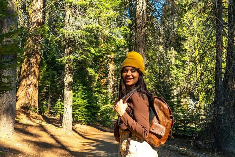 Young woman smiling and looking at camera, wearing a yellow beanie and backpack, enjoying her walking adventure on a shaded trail surrounded by tall trees in a california national park