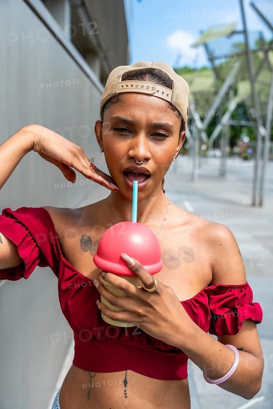 Young woman with tattoos and a cap, drinking from a unique container with a straw, showing a playful facial expression while posing in an urban street on a sunny day