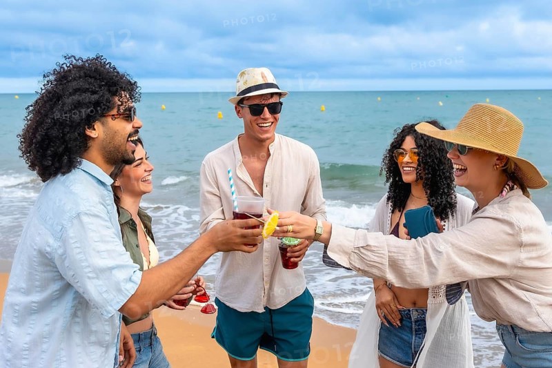 Group of diverse young adult friends cheering with colorful drinks on a sandy beach, enjoying summer vacation, feeling cheerful, and celebrating friendship with the ocean in background