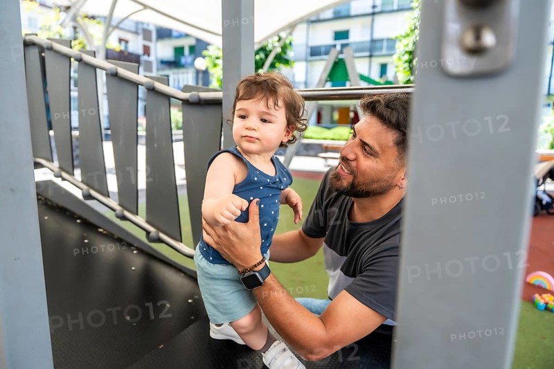 Father guiding toddler on playground structure during sunny park playtime, sharing joyful bonding, support and safety as child climbs slide in a warm, active family moment