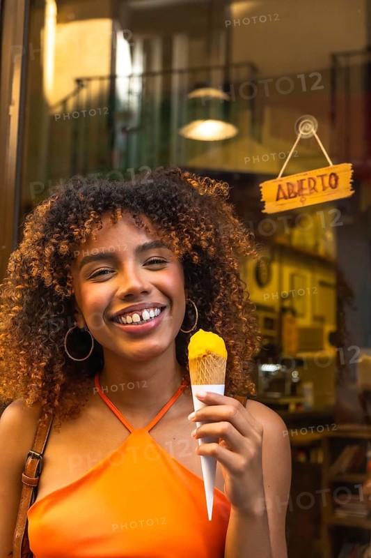 Young black woman with curly hair smiling and holding a yellow ice cream cone in front of a shop with an aberto sign, enjoying a sweet dessert outdoors