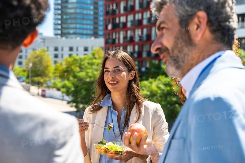 Smiling businesswoman having a healthy salad lunch and conversation with diverse colleagues outdoors during a refreshing office break in an urban setting