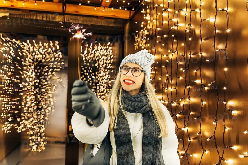 Young smiling woman holding burning sparkler while celebrating winter holidays. A person in a gray knit hat, white coat, dark scarf and gloves stands in a festive illuminated tunnel holding a lit sparkler. Warm yellow string lights fill the background with dense glowing bokeh, creating strong contrast with the winter clothing. The sparkler's bright sparks add a vivid focal point and enhance the layered warm light surrounding the figure