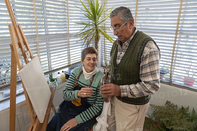 Senior couple selecting paintbrushes together beside a blank canvas in a bright home art studio