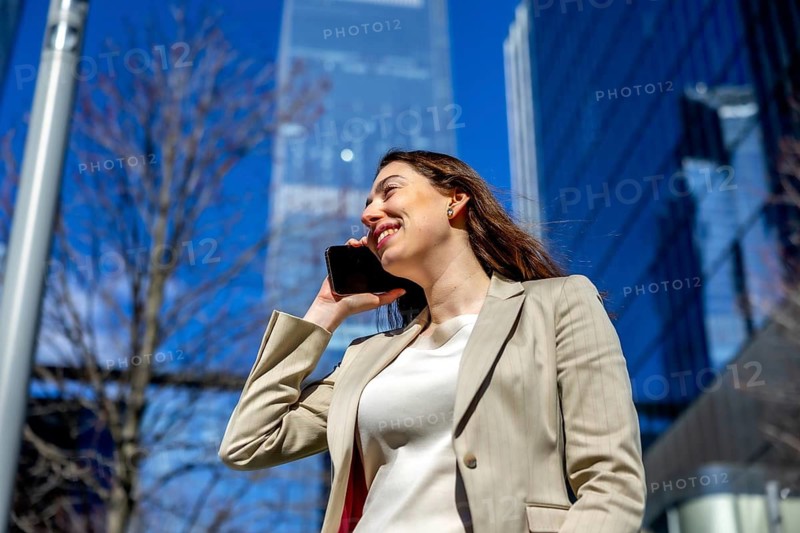 Professional young businesswoman having a smiling conversation on her smartphone in a vibrant urban environment, with modern skyscrapers reflecting the blue sky