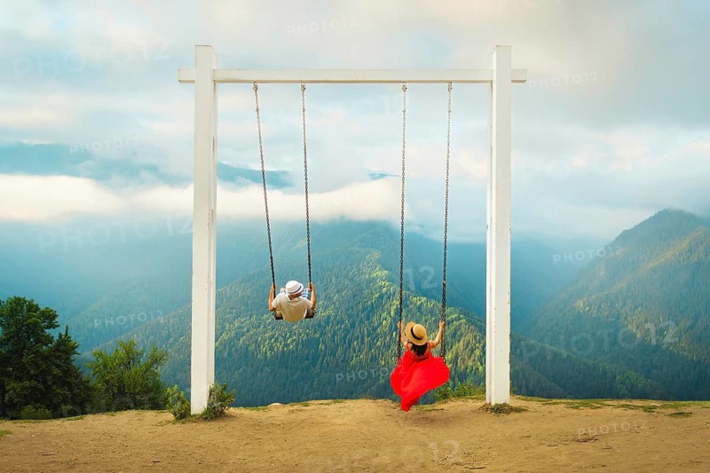 Couple riding giant swing above mountain valley with forest, clouds and panoramic landscape, symbolizing freedom, romance, adventure, wellness and dream travel