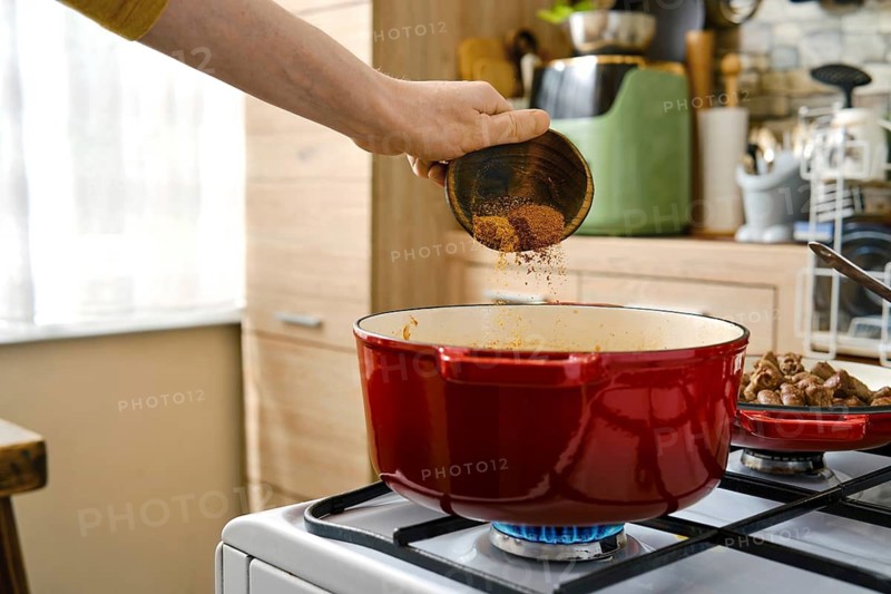A person sprinkles spices into a pot on a stove in a home kitchen. Adding flavour to a vegetable ragout