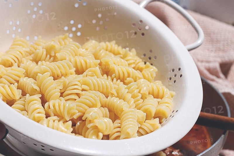 A person is holding a colander filled with cooked rotini pasta. Water is draining off as the pasta is prepared for a meal. The scene takes place in a kitchen in the afternoon