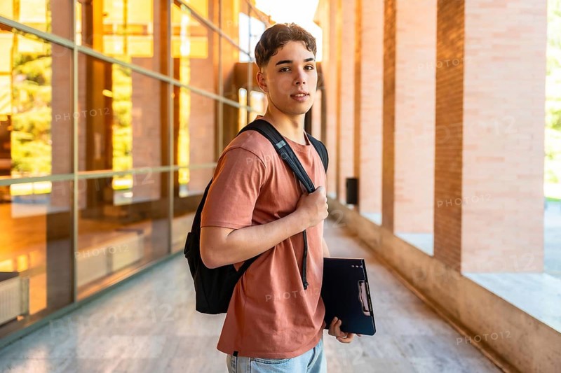 Male student walking along modern campus building with backpack and folder, reflecting college life and focused study outdoors in daytime, confident and prepared for university success
