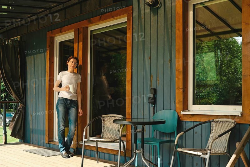 Relaxed middle-aged woman stands on the porch of wooden cabin holding drink. There are two chairs and a small table visible on the wooden deck. It is afternoon