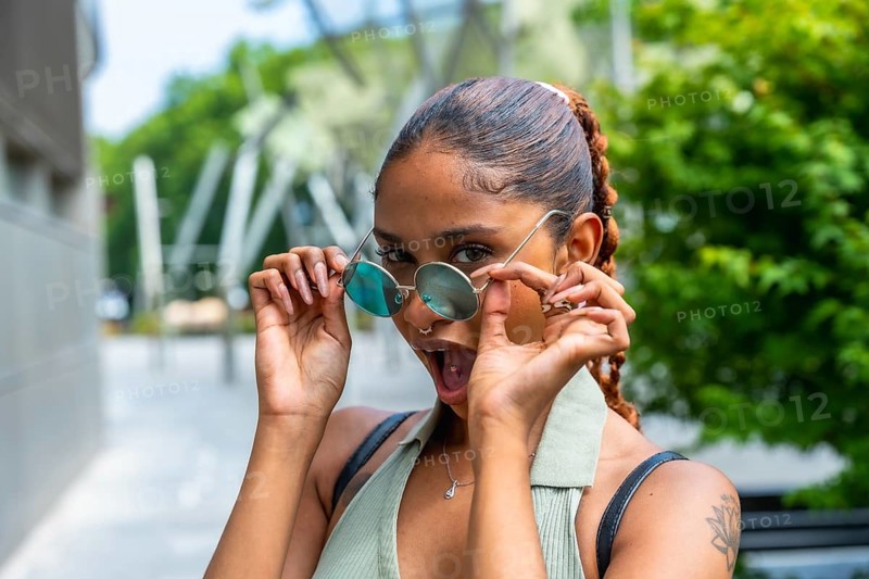 Young woman lowering round sunglasses, showing a wide open mouth expression of shock or surprise while standing on an urban street in daylight