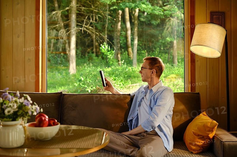 Middle-aged man sits on brown sofa in wooden cabin. He reads e-book in air-conditioned room while it is midday heat outside. Sunlight enters through window. Green trees are visible outside