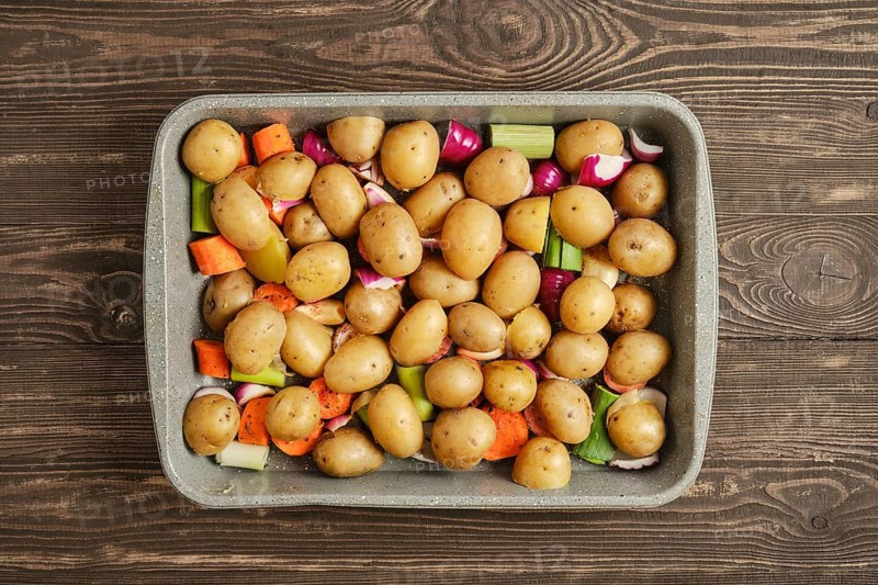 A baking dish holds an assortment of baby potatoes and chopped vegetables like carrots and celery. The dish is set on a wooden table, ready for cooking