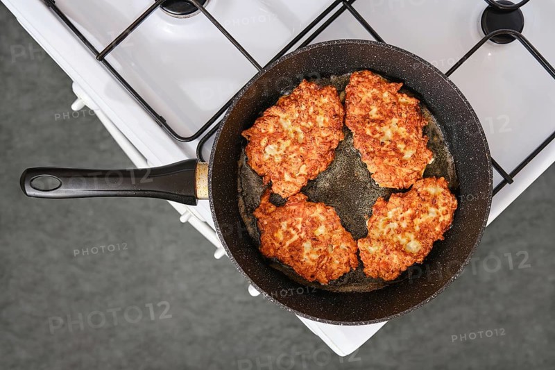 Four chicken fritters are frying in a black skillet on a gas stove. The skillet is placed on a white stove. The kitchen has a simple design with a gray floor and no visible people present