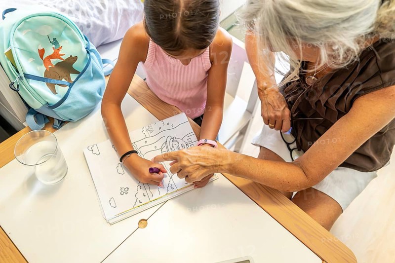 Grandmother assisting her young granddaughter coloring a book at a desk, fostering intergenerational family bonding and learning during an everyday after school activity at home