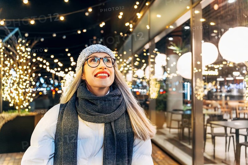 Happy young woman in winter attire under festive evening lights. A woman wearing a gray knit hat, glasses, dark scarf, gray gloves and a white winter jacket stands outdoors at night beneath warm decorative string lights. Illuminated trees and a glass-walled café with round hanging lamps create a bright festive holiday atmosphere. The combination of winter attire, glowing urban surroundings and cozy seasonal lighting conveys a strong Christmas and New Year mood