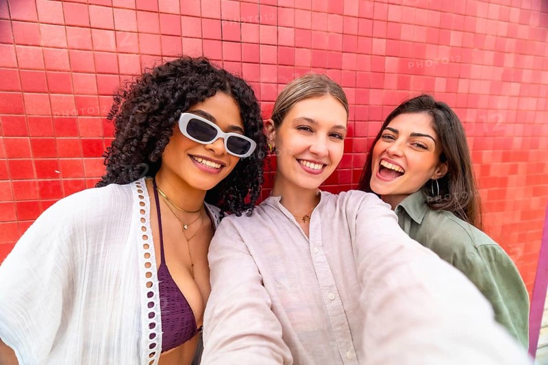 Diverse young women smiling and laughing while taking a summer selfie together against a vibrant red wall, enjoying friendship, sunshine and carefree urban fun