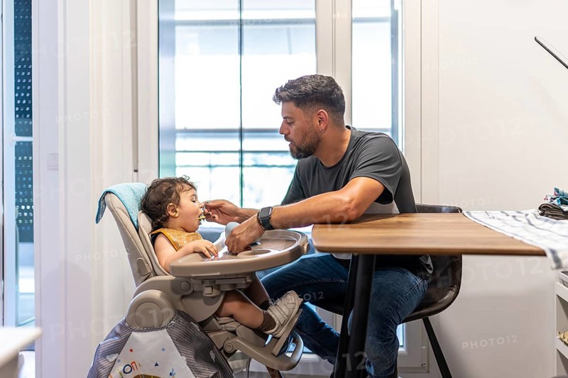 Father feeding his baby son pureed food in a high chair at home, nurturing bonding and healthy mealtime habits in a warm, modern family kitchen setting