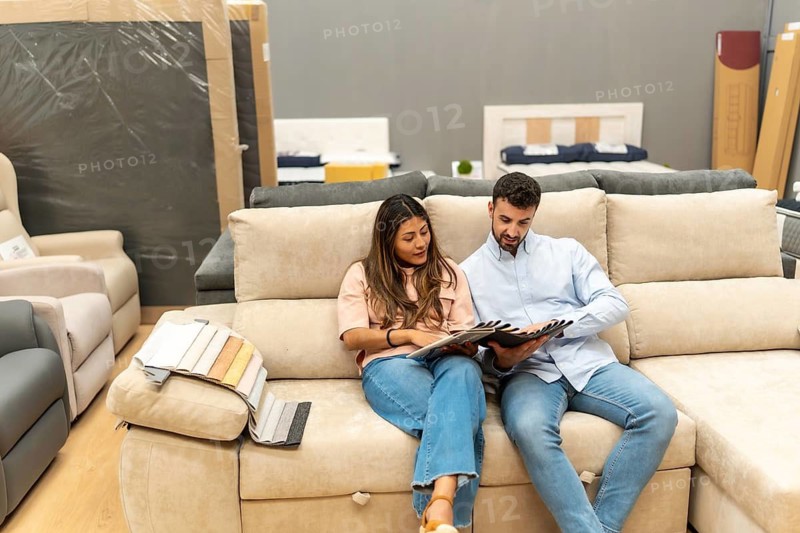 Young couple sitting on a large sofa, carefully examining different fabric samples and discussing options for home decoration in a modern furniture showroom, making purchase decisions