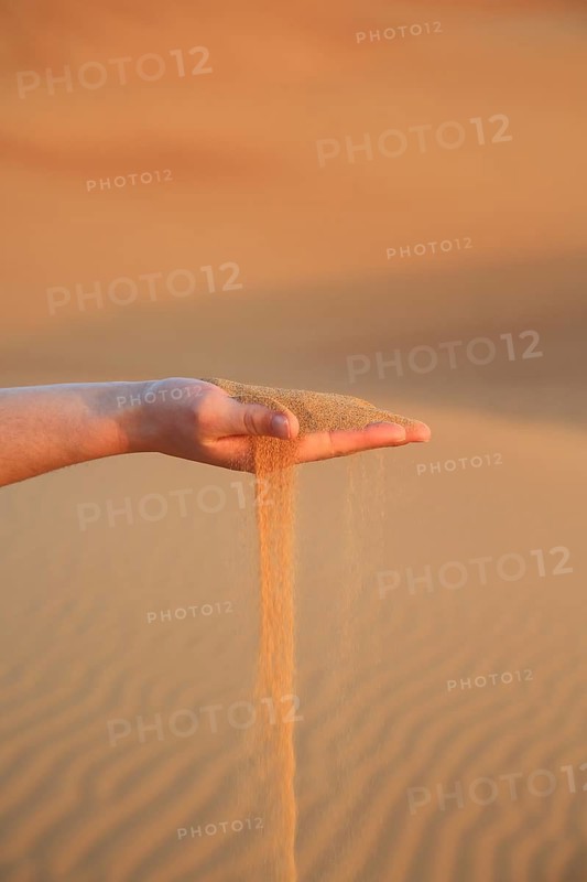 Hand holding trickling sand in a quiet desert landscape at sunset, A woman's hand lets red desert sand fall through her fingers and shows the connection with nature in the landscape, desert, sunrise, Dubai, United Arab Emirates