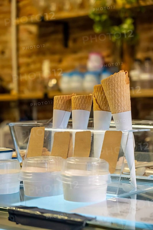Waffle ice cream cones and disposable cups standing on a illuminated glass shelf in a dessert shop, ready for serving gelato or sweet treats to customers