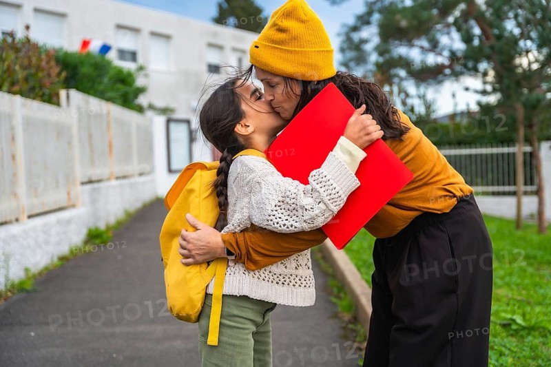 Loving mother kisses and hugs her young daughter on the sidewalk as she heads to school, tender morning routine capturing family bond, support and back to school emotion