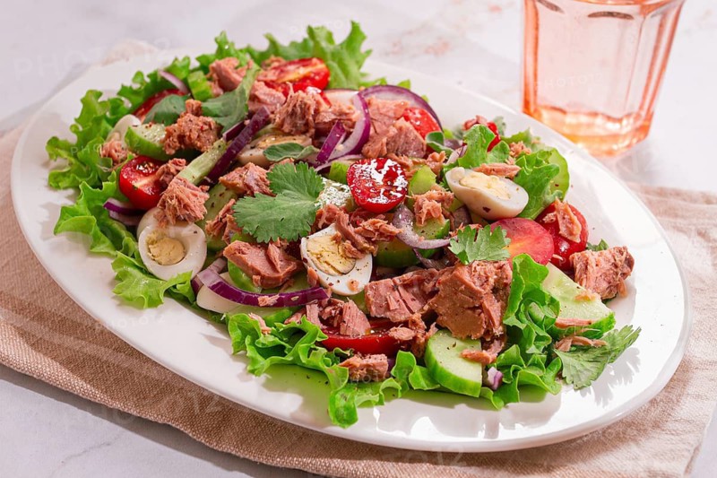 A fresh salad is served on a white platter. The salad includes tuna, cherry tomatoes, cucumber, boiled eggs, and lettuce