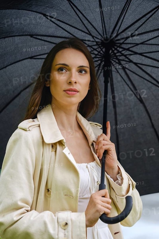 Woman stands outside with black umbrella in hand. She is wearing a light coat and looks thoughtful