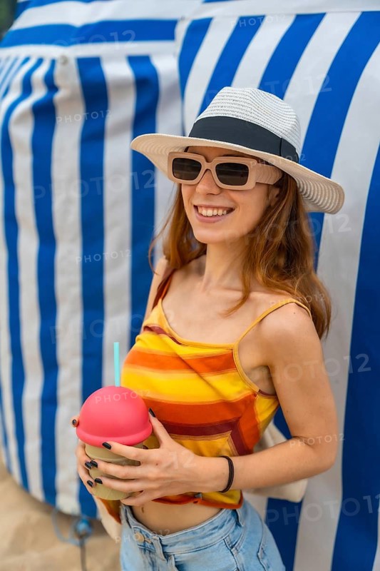 Young woman wearing a hat and sunglasses, smiling while enjoying a cold beverage on a sunny summer day, standing in front of blue and white striped fabric