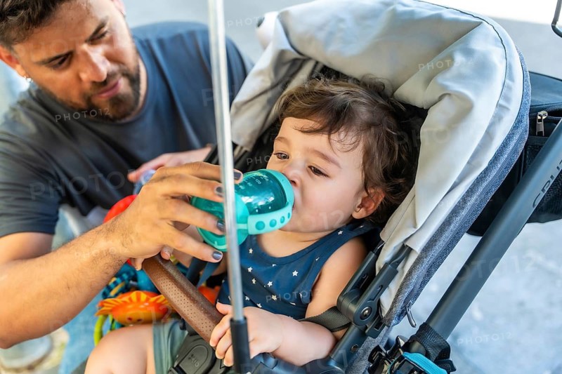 Father offering a sippy cup of water to his toddler son in a stroller during a warm urban stroll, keeping him hydrated and comfortable while bonding outdoors