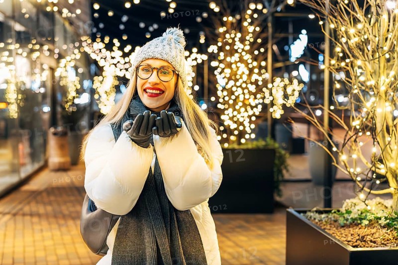 Young woman in winter attire enjoying festive lights outdoors. A woman wearing a white winter coat, gray knit hat with a pom-pom, dark scarf, gray gloves and glasses stands outdoors at night, smiling with her hands near her face. Warm white string lights wrap the nearby trees, creating a bright festive glow. Glass storefronts and the urban walkway add depth to the cozy Christmas and New Year atmosphere