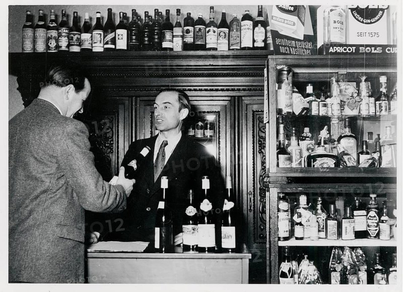 West Berlin, shop for alcoholic beverages, pub, A bartender serves a man in front of a shelf full of bottles, around 1960