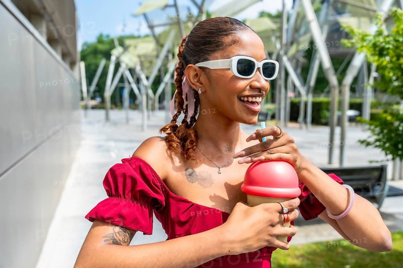 Young woman with braided hair and sunglasses, wearing a red off shoulder dress, smiling and holding a refreshing cold drink with a straw in a sunny modern city environment