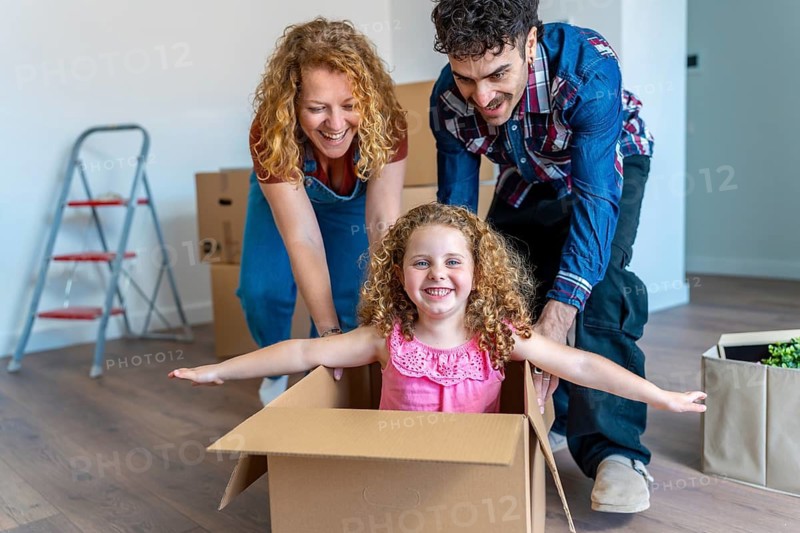 Parents pushing their young daughter in a cardboard box on a wooden floor, celebrating their relocation to a new home and creating joyful memories during the moving process