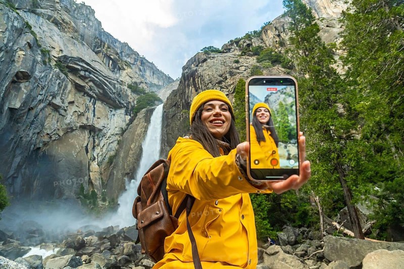 Young woman in yellow raincoat holding a smart phone, recording a selfie video while visiting lower yosemite falls in yosemite national park, embracing nature and technology