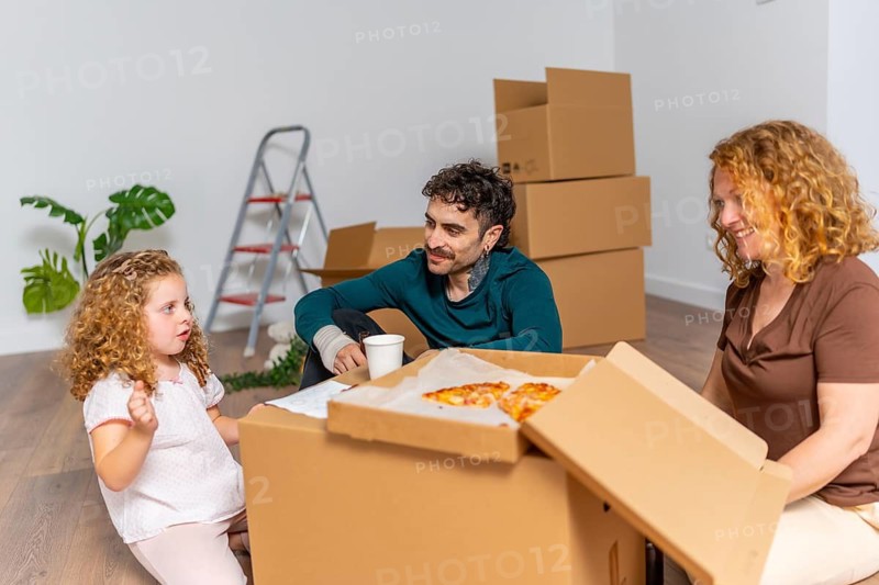 Happy family with a young daughter relaxing on the floor, eating pizza from a cardboard box, and celebrating their new home surrounded by unpacked moving boxes, signifying a fresh start